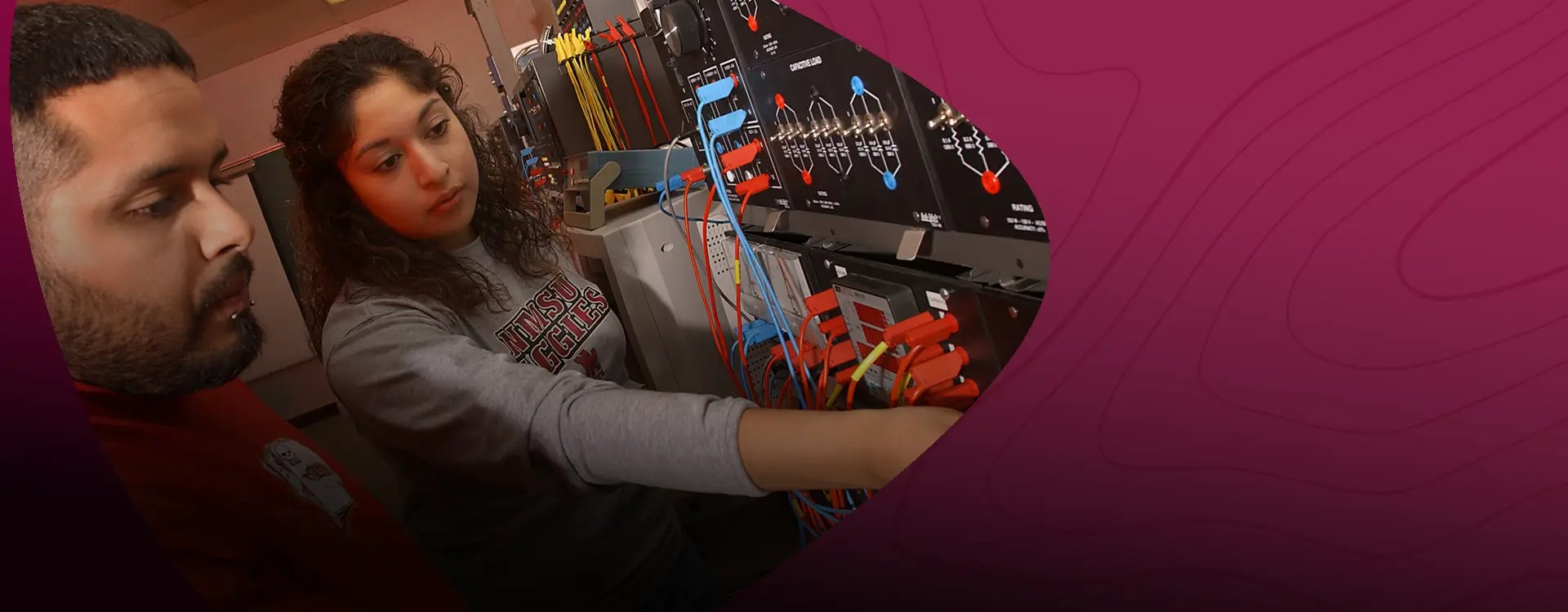 A young woman in an NMSU Aggies sweatshirt examines a technical control panel with her coworker— NMSU Global Campus